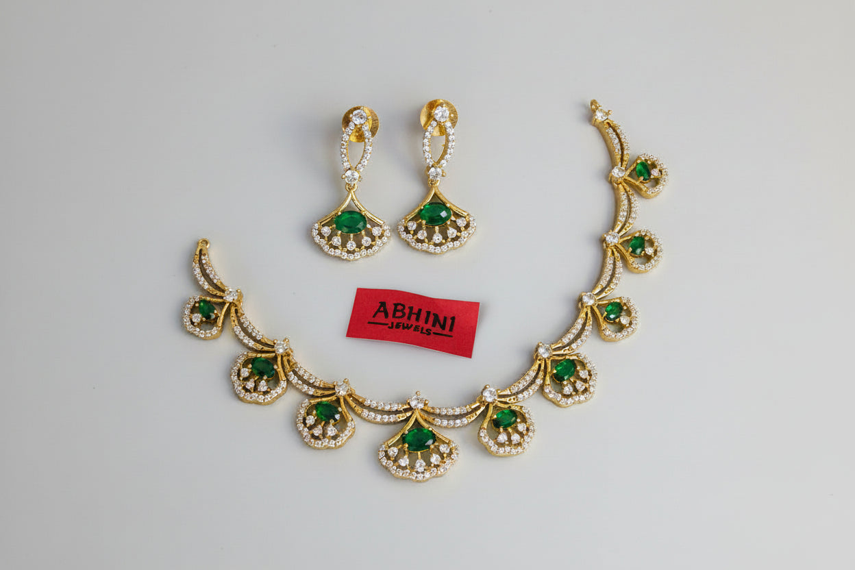 Gold necklace with green stones and matching earrings on a gray background, branded 'Abhini'.